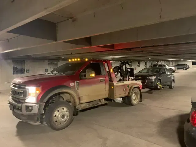 A red tow truck towing a gray car in a parking garage. The truck is dirty.