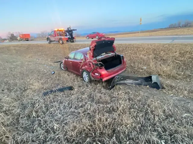 Red car, heavily damaged in a crash, off the side of the road with tow truck in the background.
