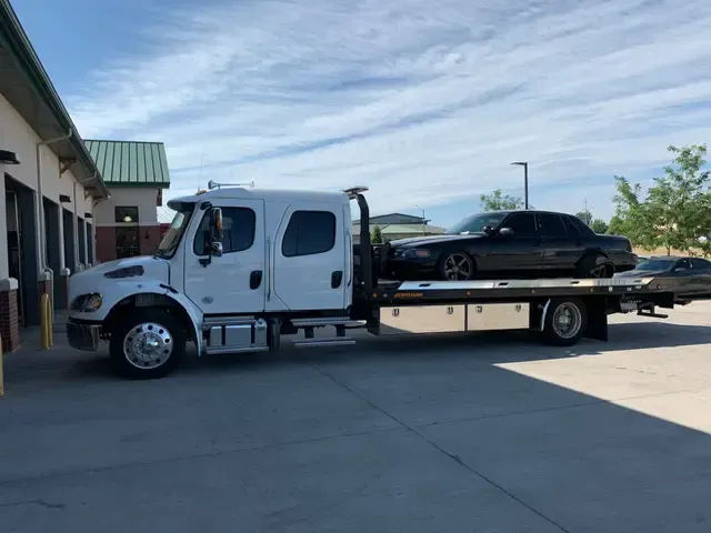 White tow truck with black sedan on its flatbed, parked outside a building on a sunny day.