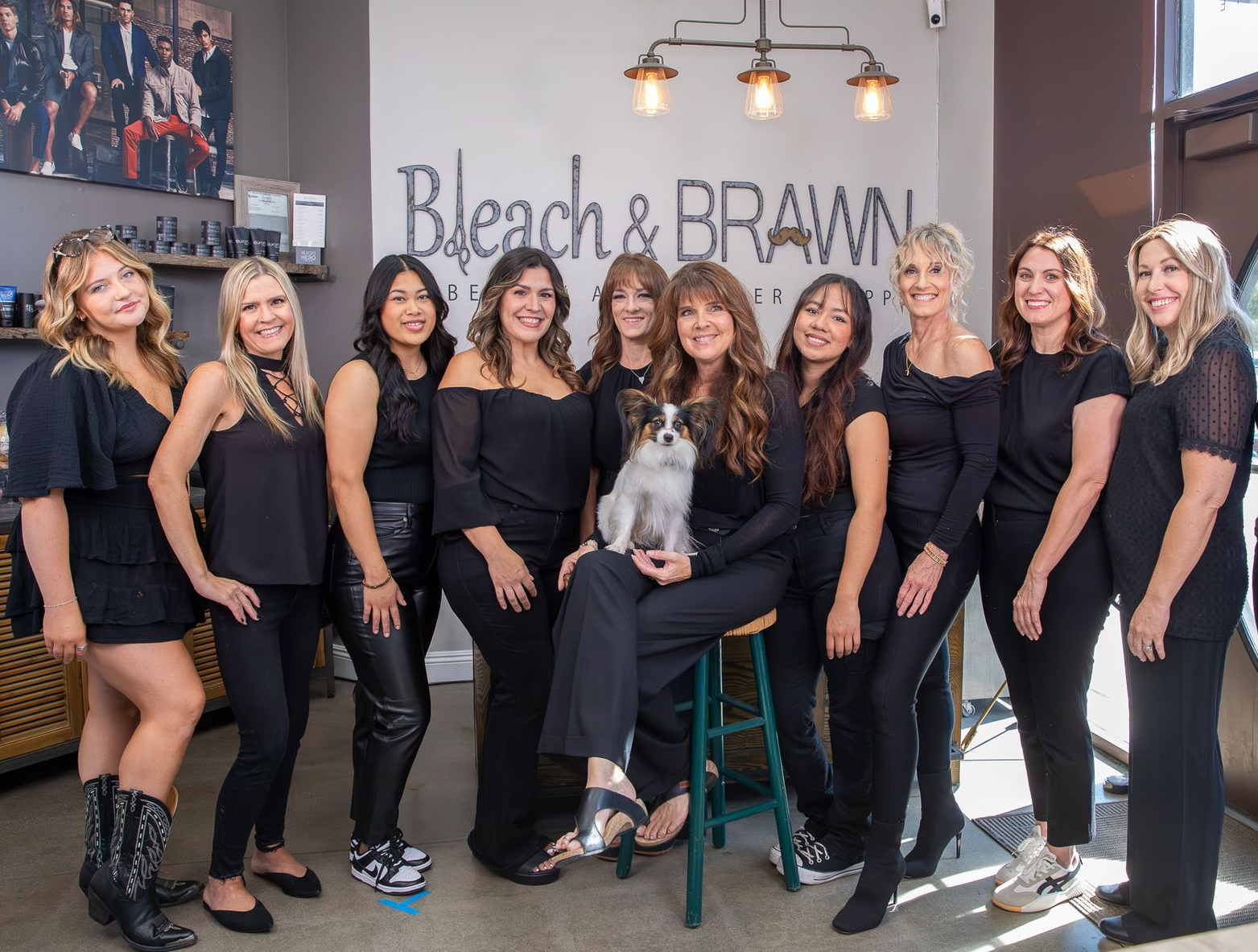 Group of women in black clothing posing in a hair salon. A small dog sits in front. 