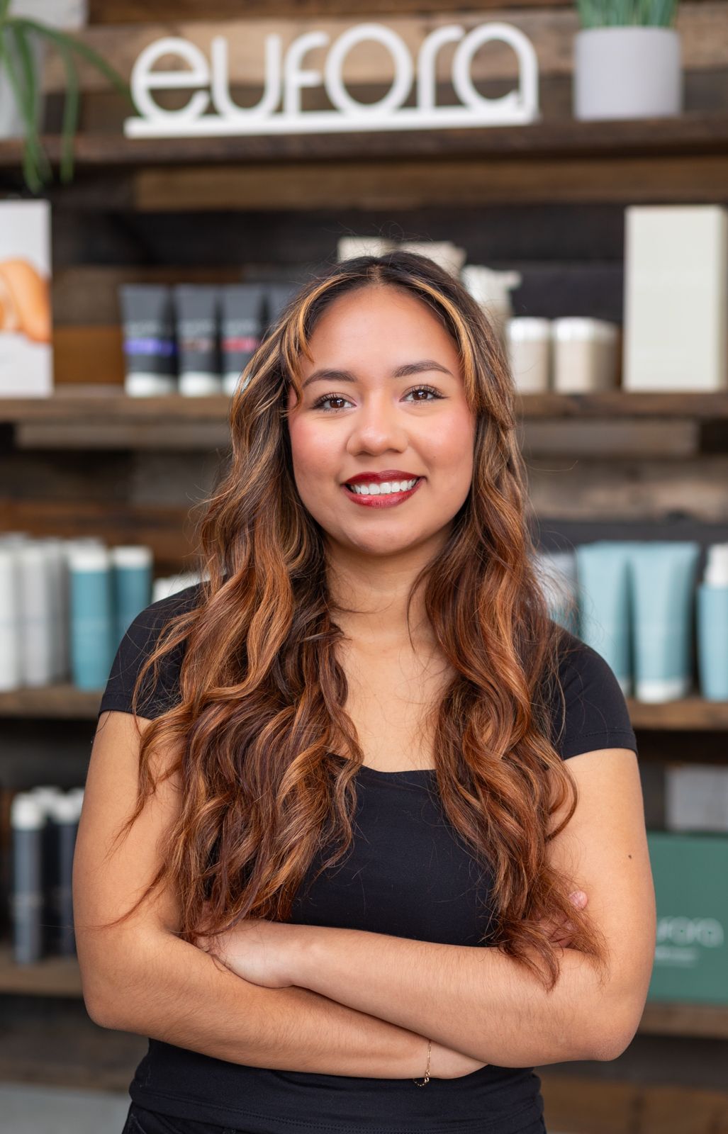 Woman smiles, leans on wooden counter. She wears a black shirt and pants, against a store backdrop.