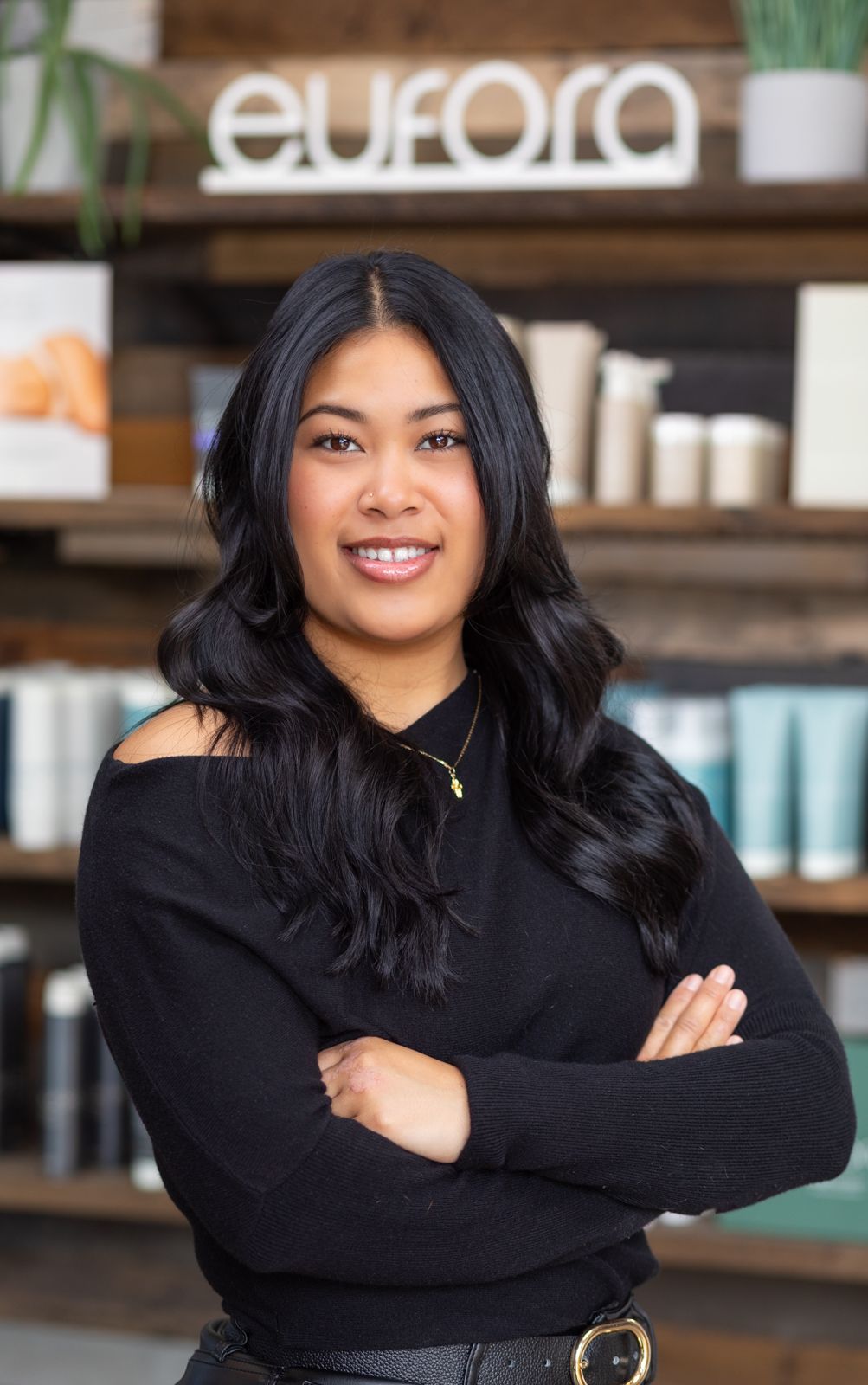 Woman leaning on a wooden counter, smiling. Wearing a black top and leather pants. Background blurred.