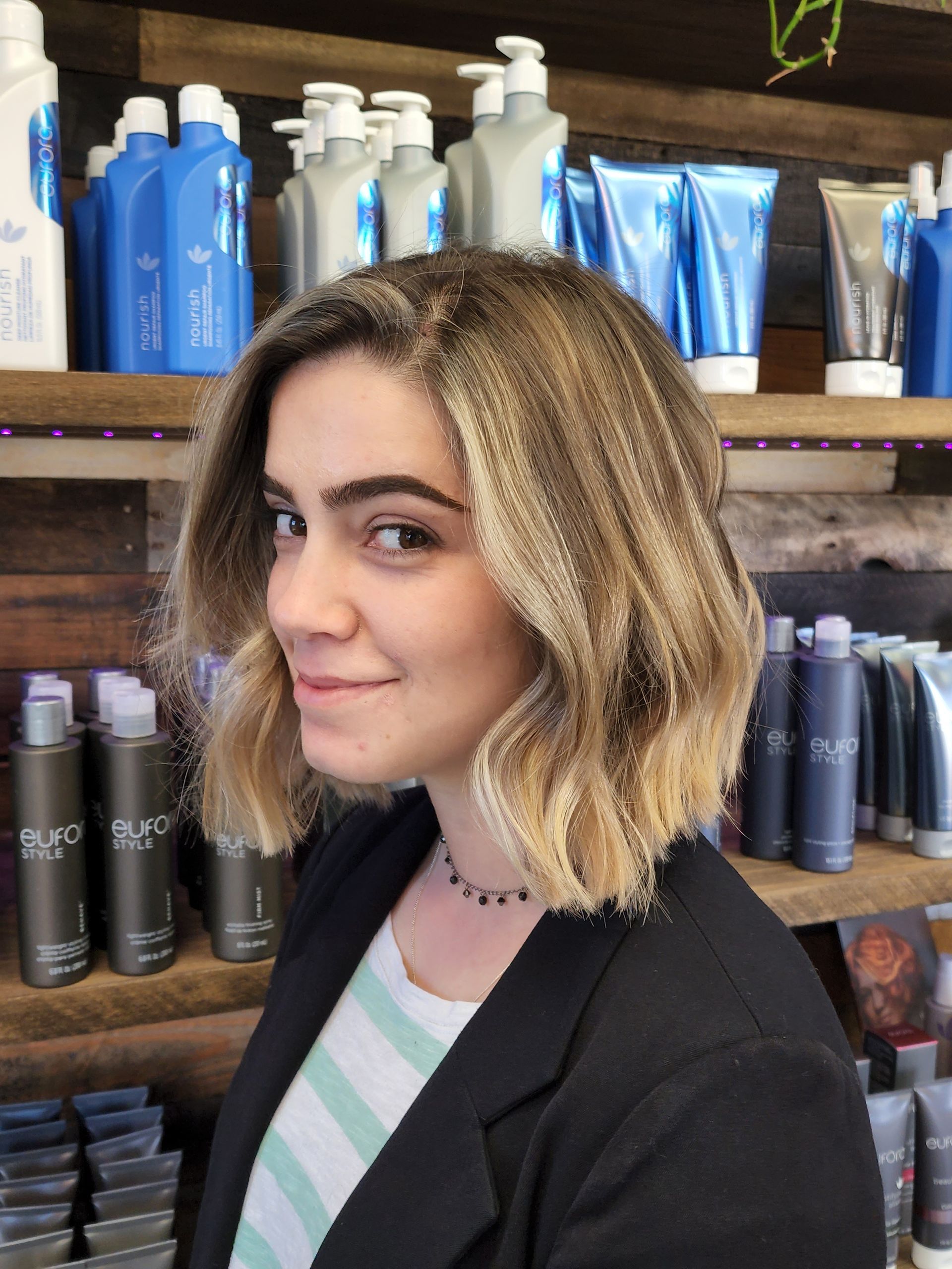 Woman with wavy blonde hair smiles, standing in front of a shelf with hair products.
