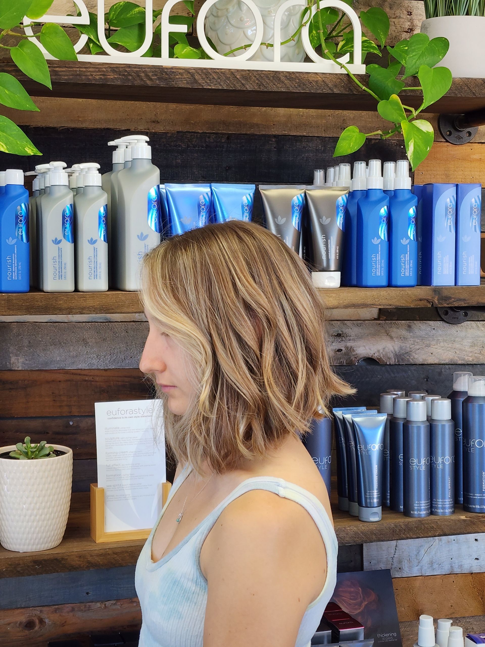 Woman with crimped, shoulder-length blonde hair stands in a salon. Shelves of products behind her.