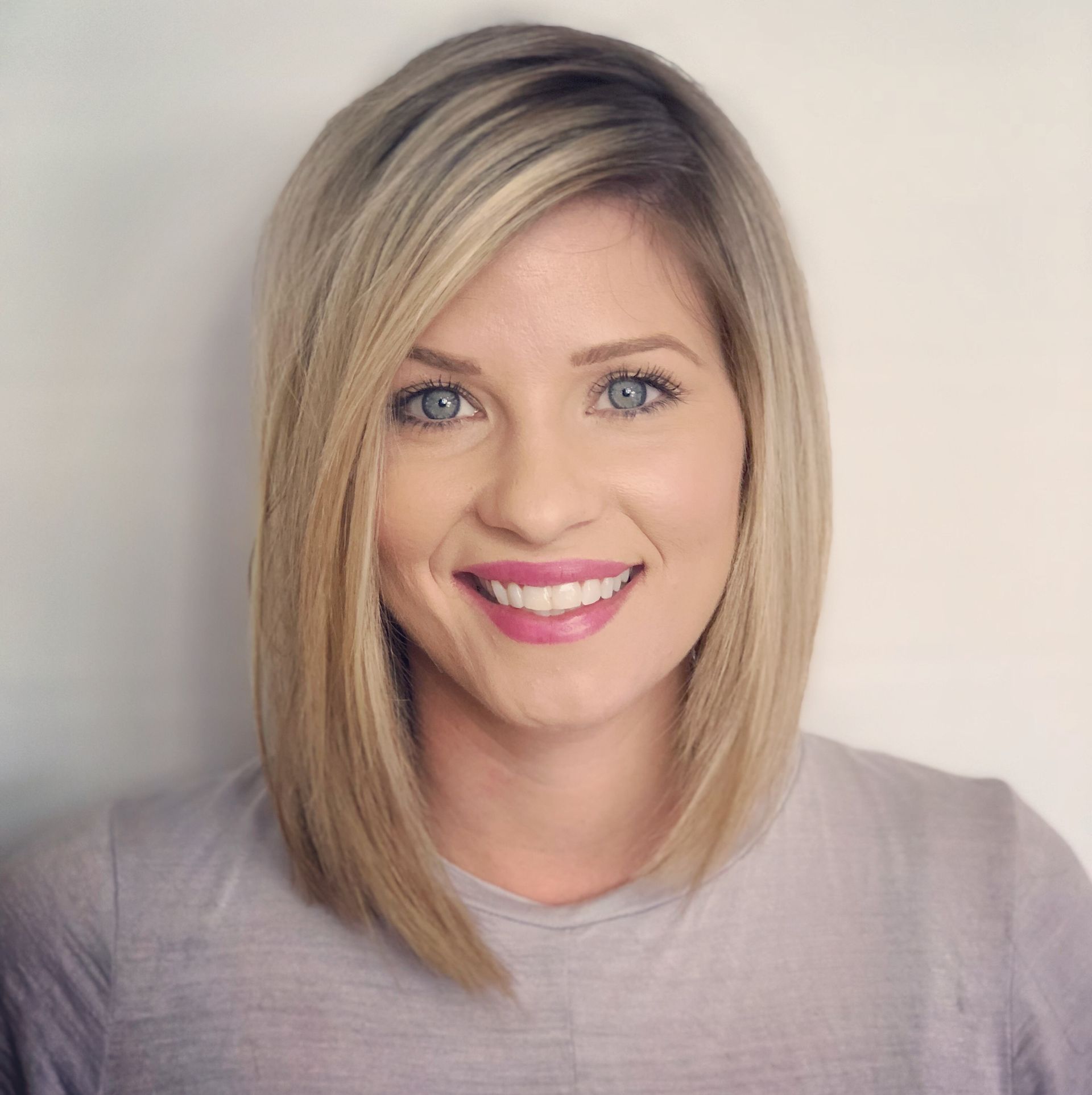 Blond woman with blue eyes smiles, wearing a gray shirt, against a white backdrop.