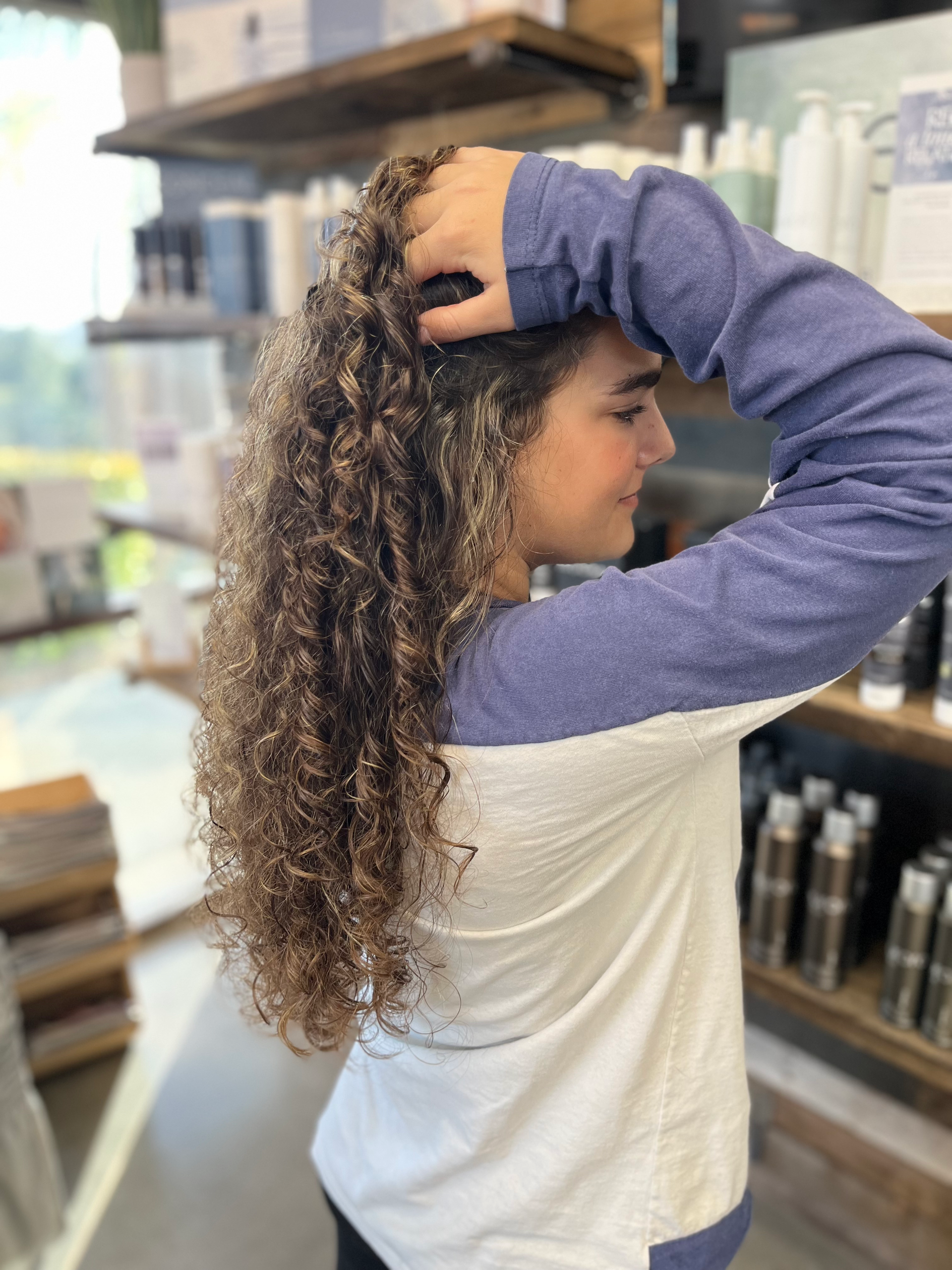 Woman with long, curly hair touches it with one hand. Wearing a blue and white long-sleeved shirt, in a shop.
