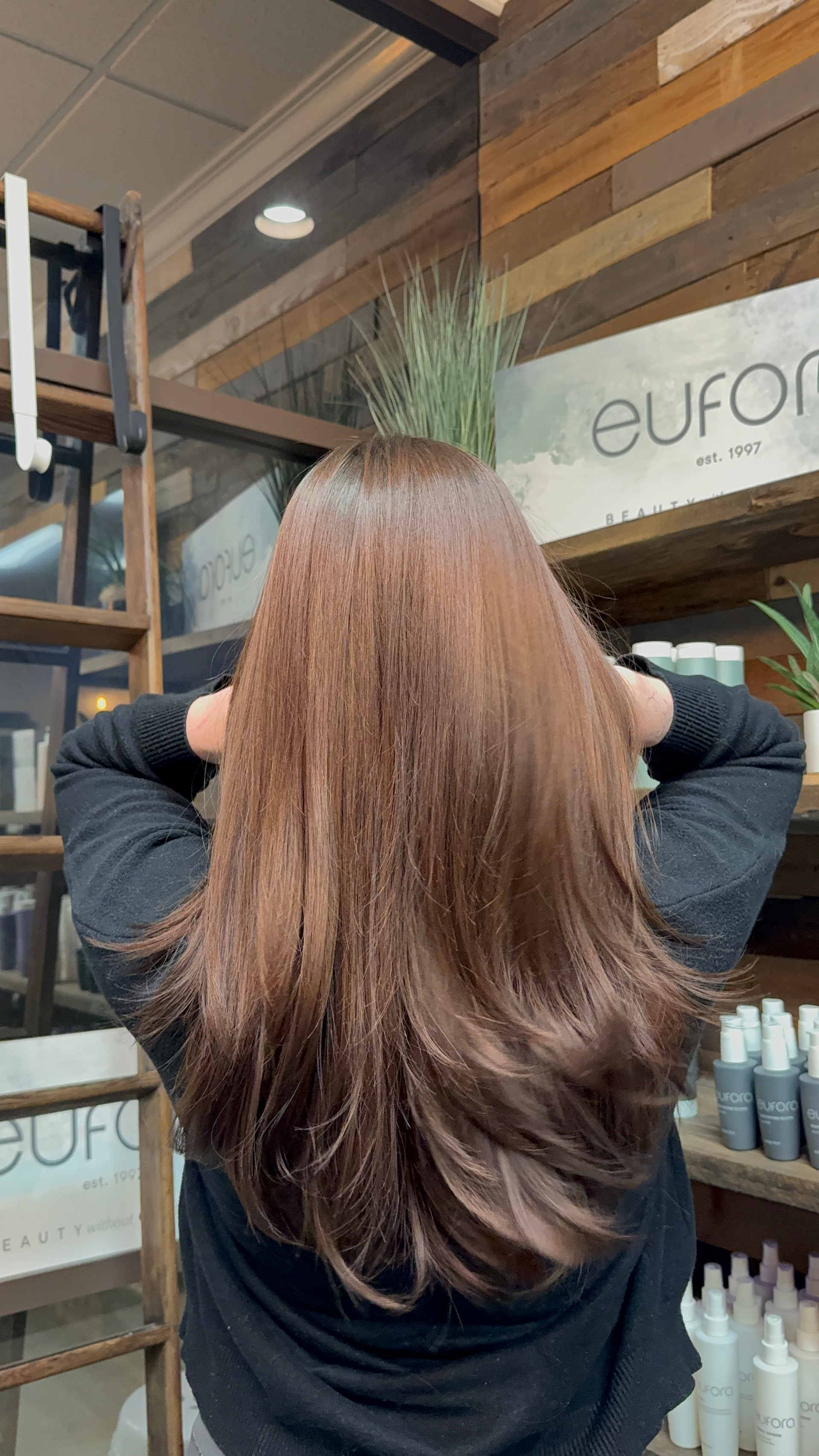 Woman with long, shiny, brown hair, facing away, hands behind her head. Salon background with product display.