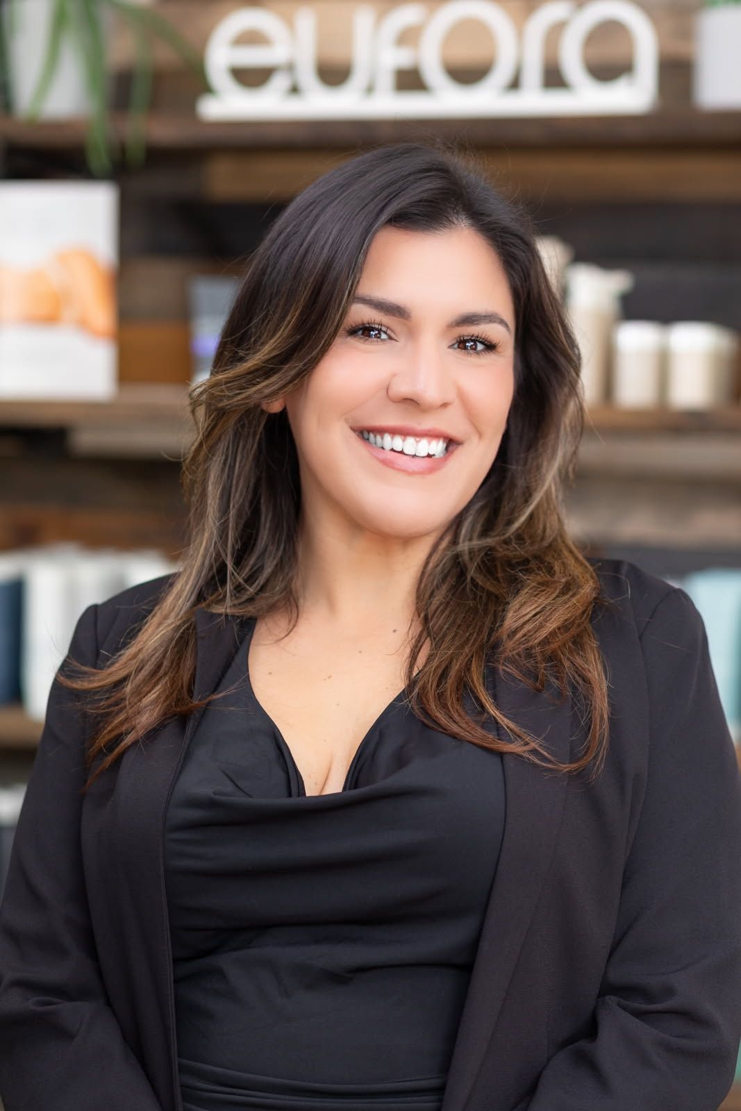 Woman smiling, wearing black off-the-shoulder top, leaning on wooden counter; in front of a shop sign.