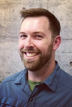 Smiling man with short brown hair, beard, wearing blue shirt and green undershirt, against a textured gray background.