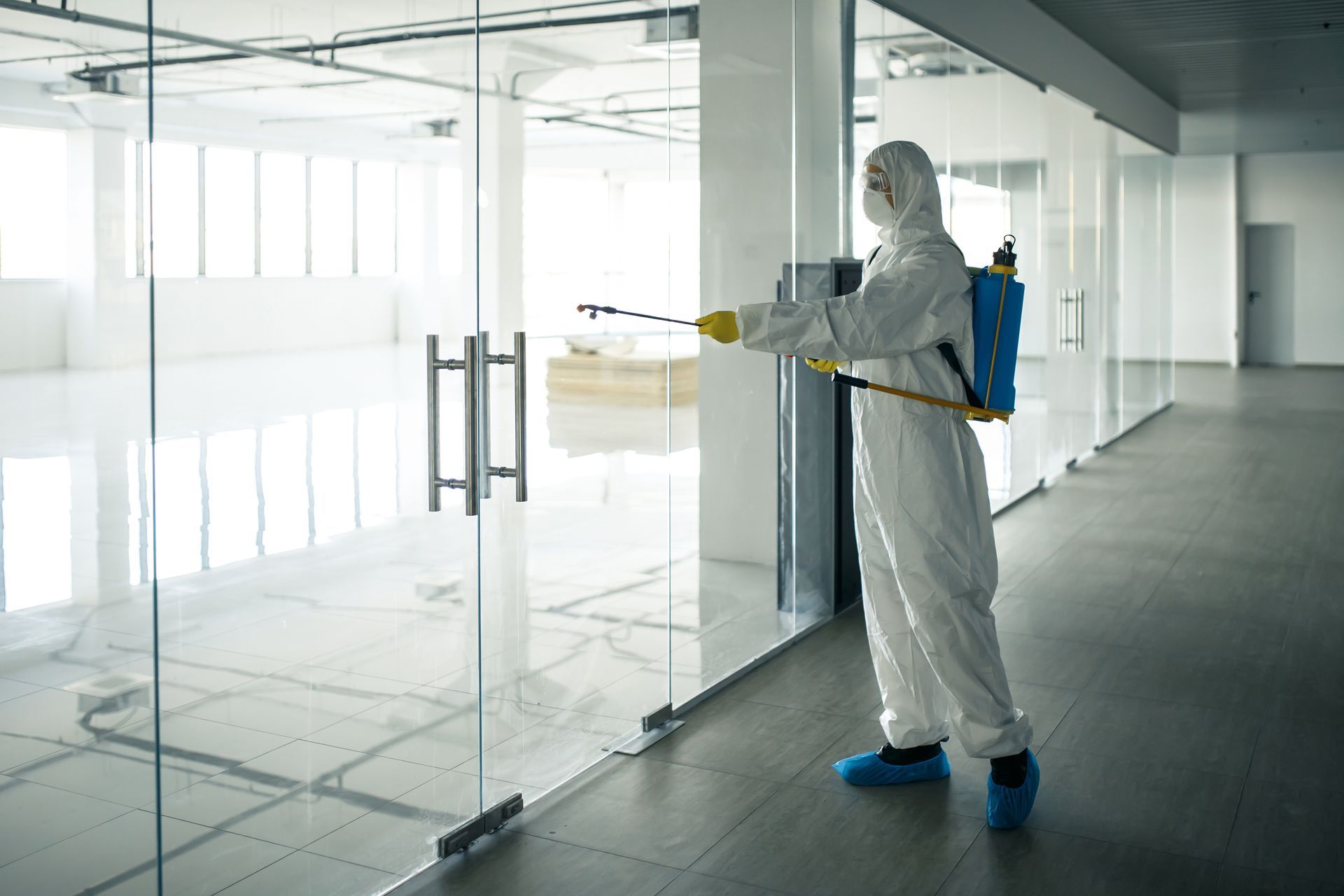 A man in a protective suit is spraying disinfectant on a glass door.