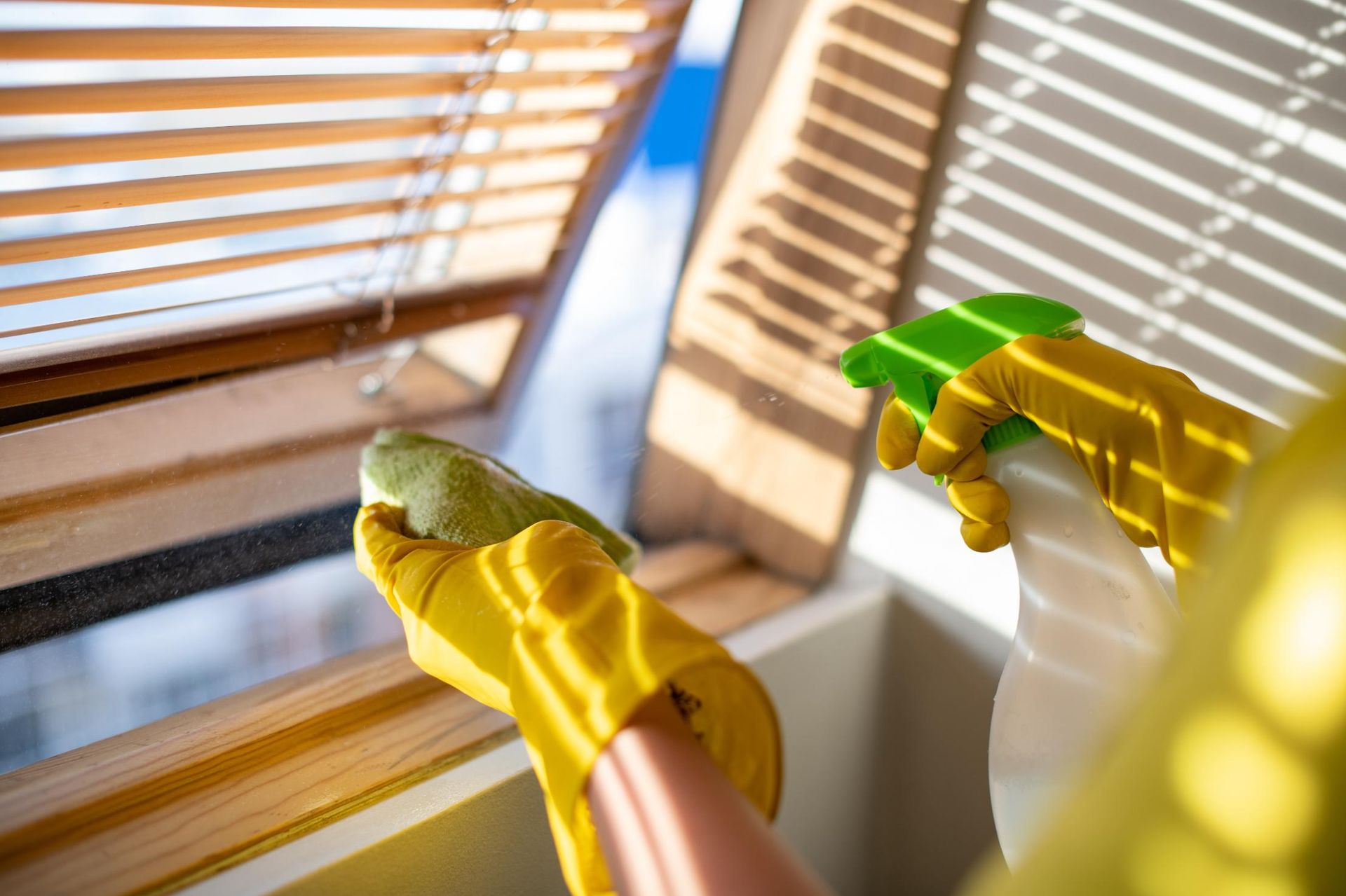 A person wearing yellow gloves is cleaning a window with a cloth and spray bottle.