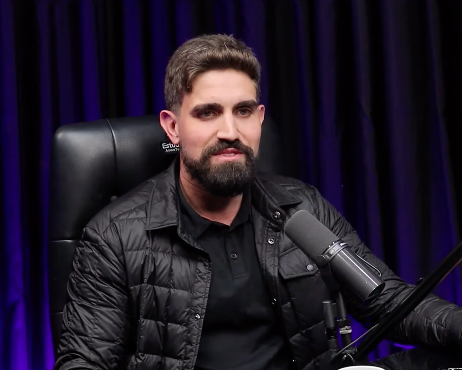 Um homem com barba está sentado em uma cadeira em frente a um microfone.