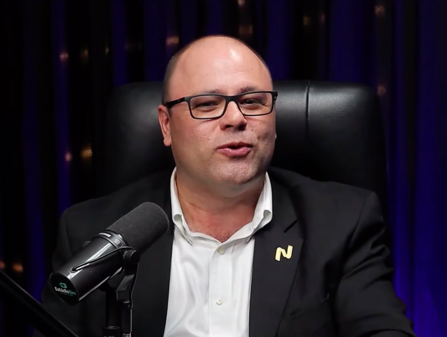 Um homem de terno e óculos está sentado em frente a um microfone.