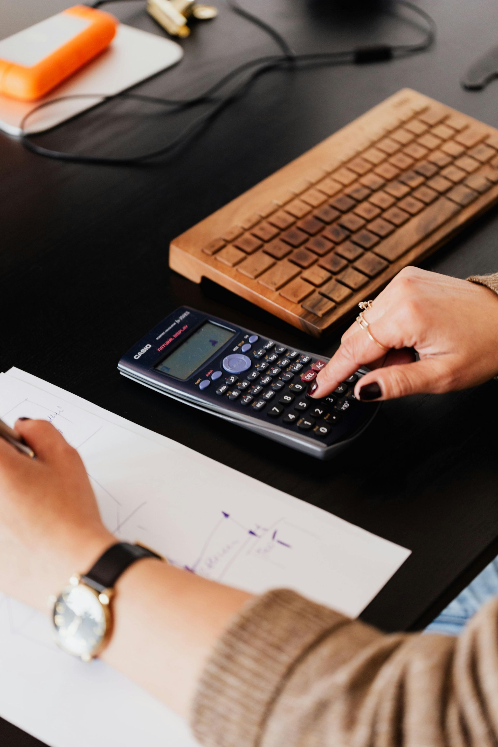 Person using a scientific calculator on a black desk, with a wooden keyboard, paperwork, and an external hard drive visible.