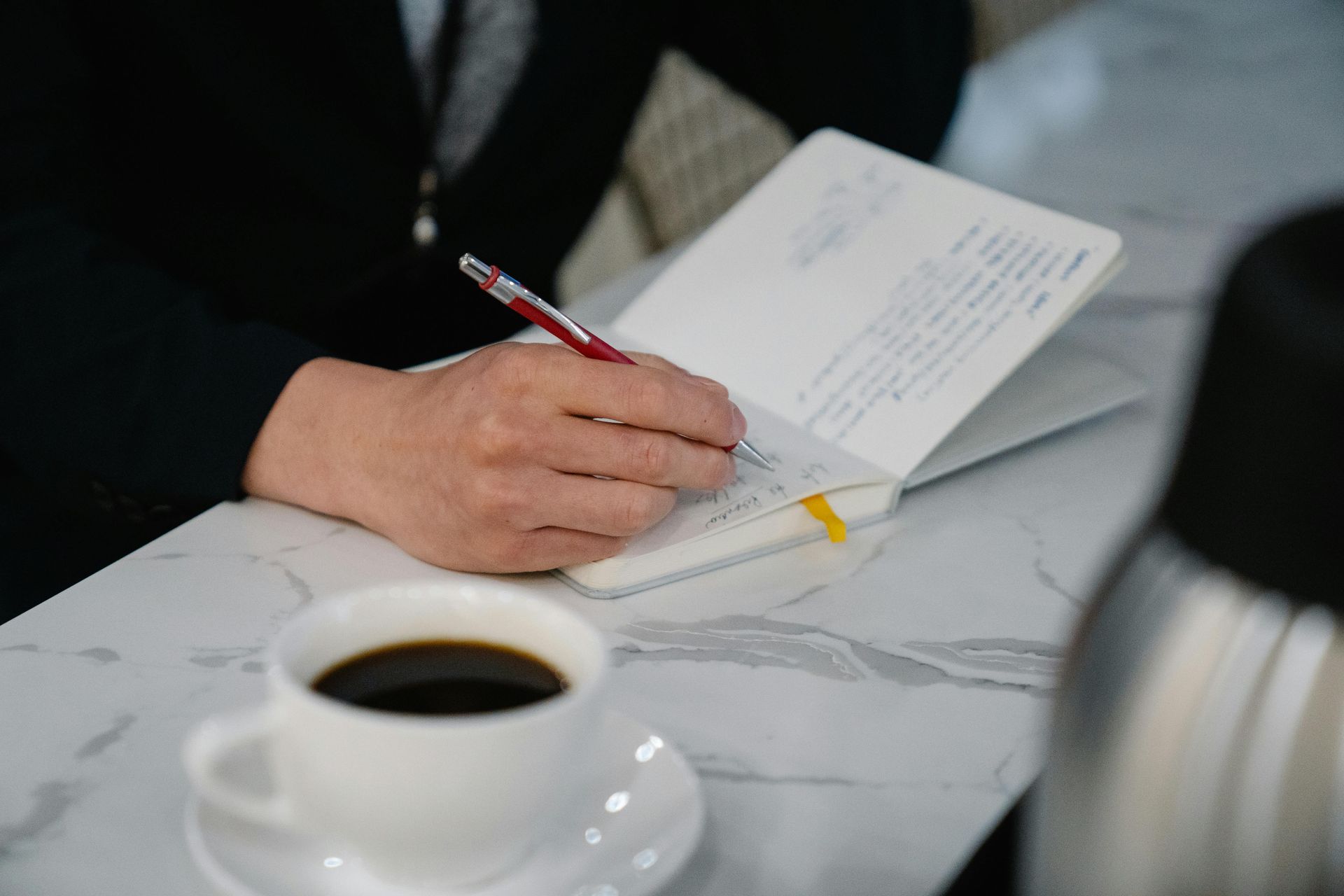 Person in a black suit writes in a notebook with a red pen next to a cup of black coffee on a white table.