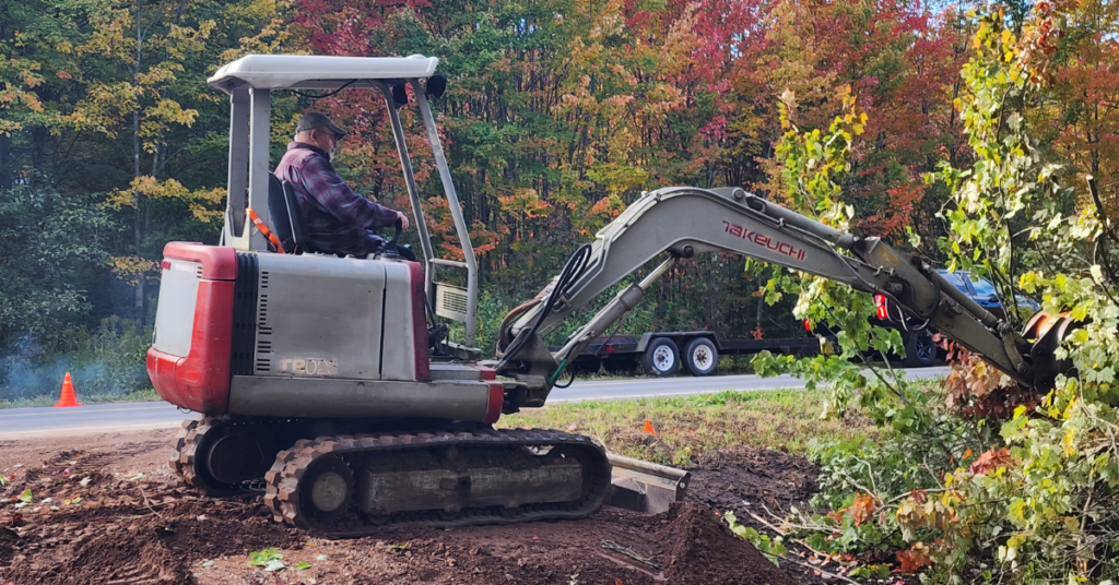 A person operates a small excavator, trimming bushes beside a road, fall foliage in the background.