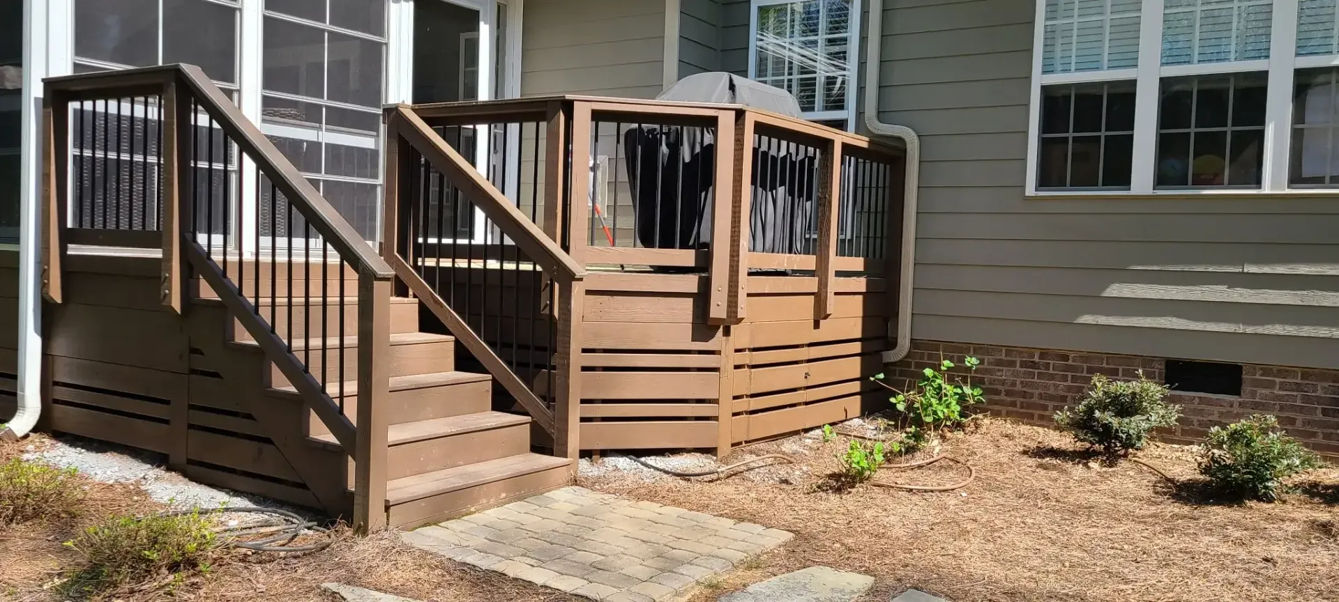 A wooden deck with stairs and railing is built against a house with windows. The yard is covered in wood chips.