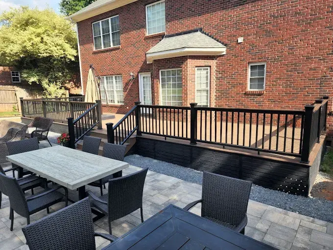 A brick house with a deck and patio. Black railing encloses the deck. A stone patio has outdoor dining furniture.