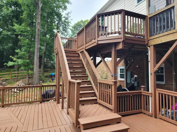 A wooden deck with stairs leading to an elevated platform. Brown railings and steps, with a forest background.