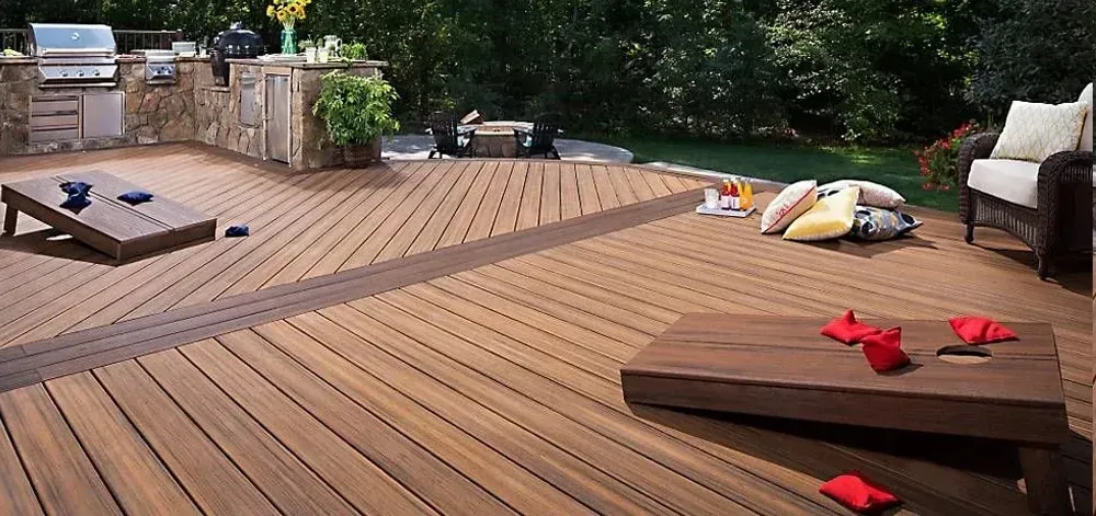 A wooden deck with two cornhole game boards set up. Backyard patio with a built-in grill and lush green foliage.