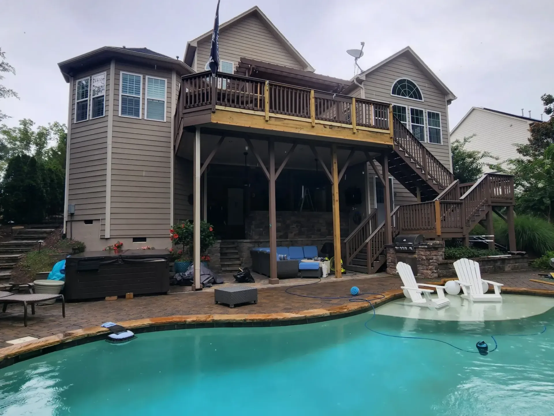 A multi-level deck and house overlook a swimming pool.  Wooden deck with steps, columns, and a sitting area. White Adirondack chairs near the pool.