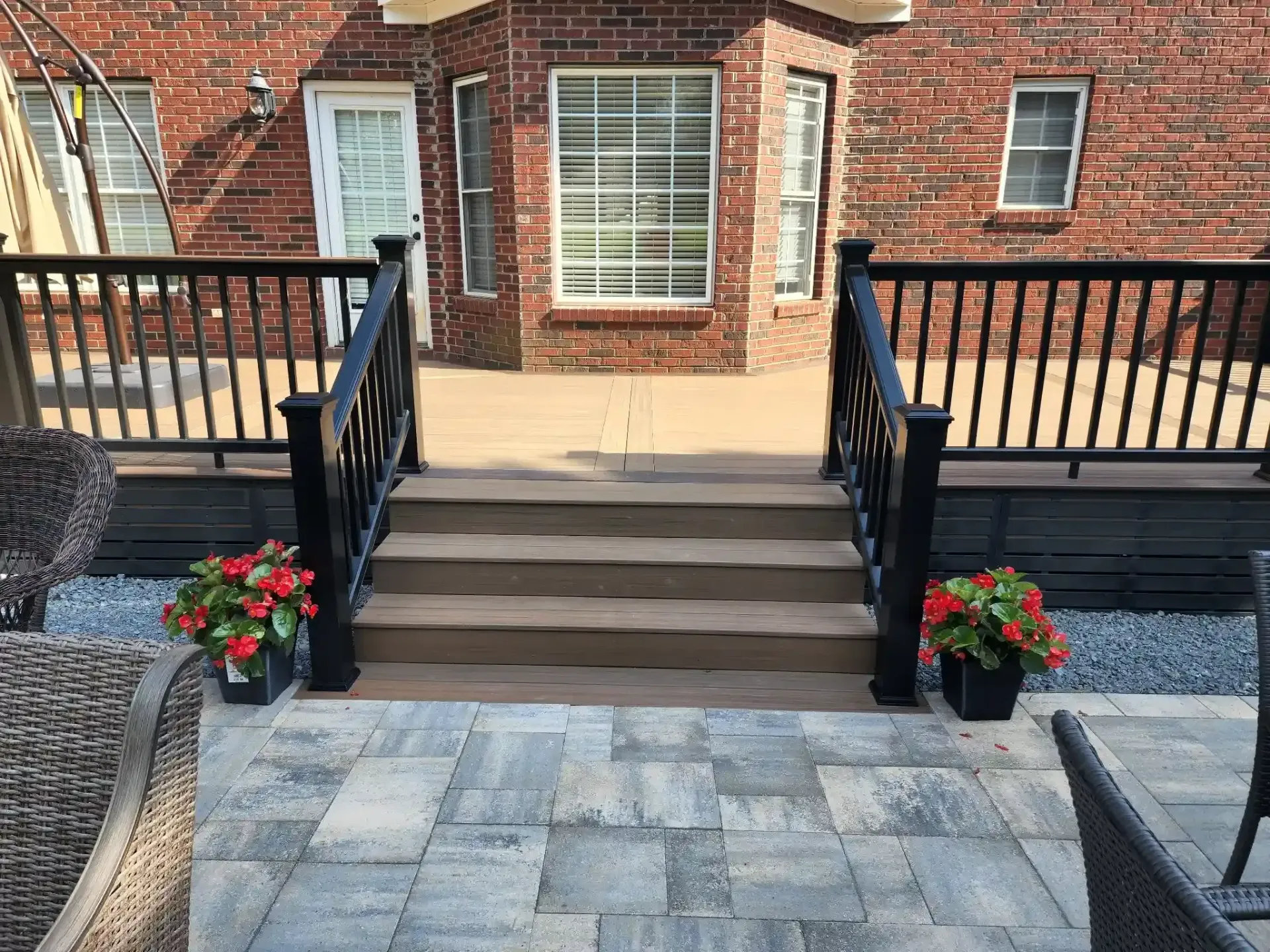 A brick house with a wooden deck accessed by steps, framed by black railings. Potted flowers flank the steps, and a stone patio sits in the foreground.