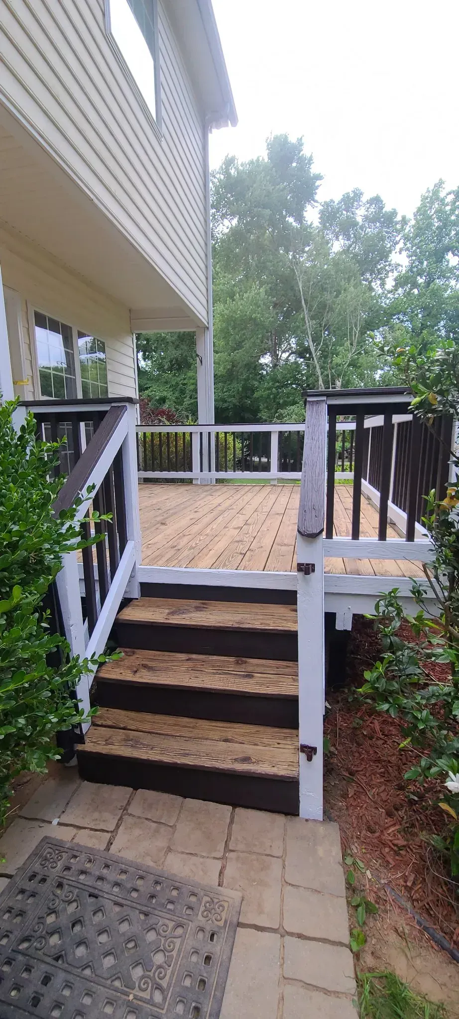 Wooden deck with stairs leading to it, beside a house. Dark stained railings and steps, white supports. Green bushes surround the deck.