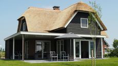 Cottage with thatched roof and dark wooden exterior, with a covered patio area, set in a grassy yard under a blue sky.