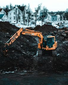 Yellow excavator working on a construction site, with houses in the background. It is likely a cloudy day.