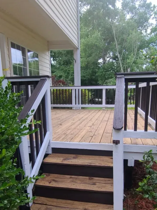 Wooden deck with brown railings, accessed by stairs. The deck is attached to a beige house and surrounded by lush greenery.