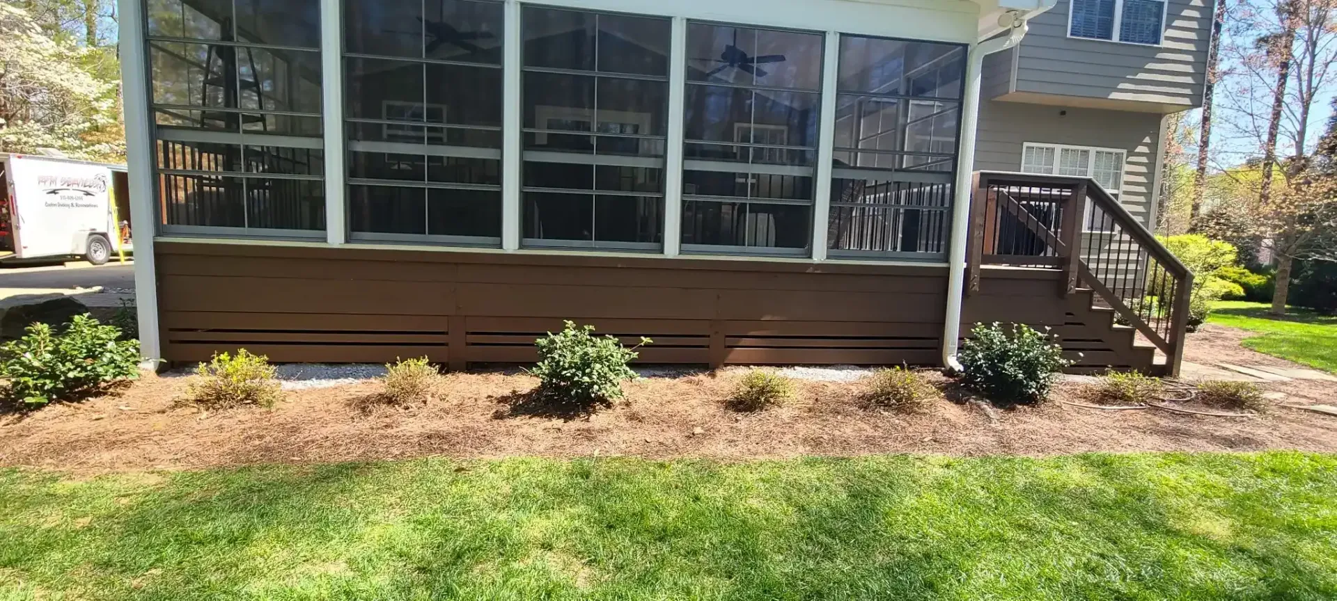 A screened porch with brown siding and a dark wooden deck, set on a bed of mulch with some shrubs in front, and green grass below.