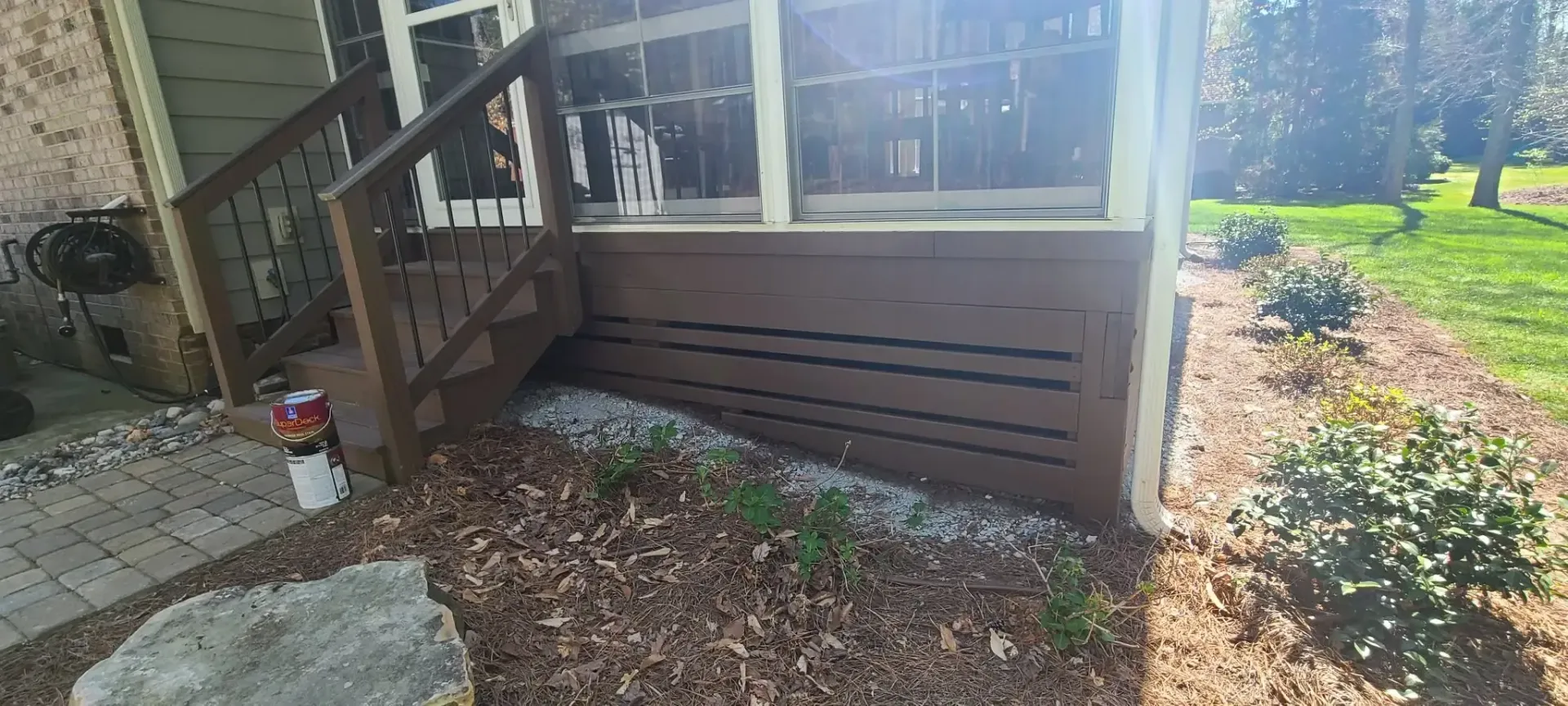 A brown wooden porch with a staircase and a flower bed in front of it. The ground is covered with brown leaves and small plants.