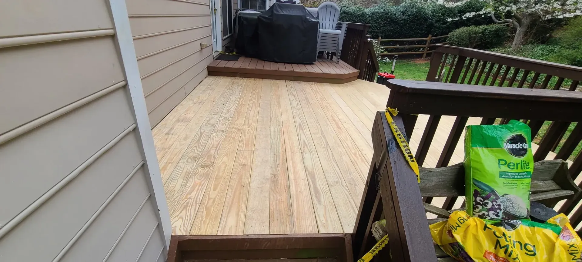 A wooden deck with freshly laid planks, surrounded by a beige siding wall on one side and a railing with gardening supplies on the other, with green grass and a wooden fence in the background.