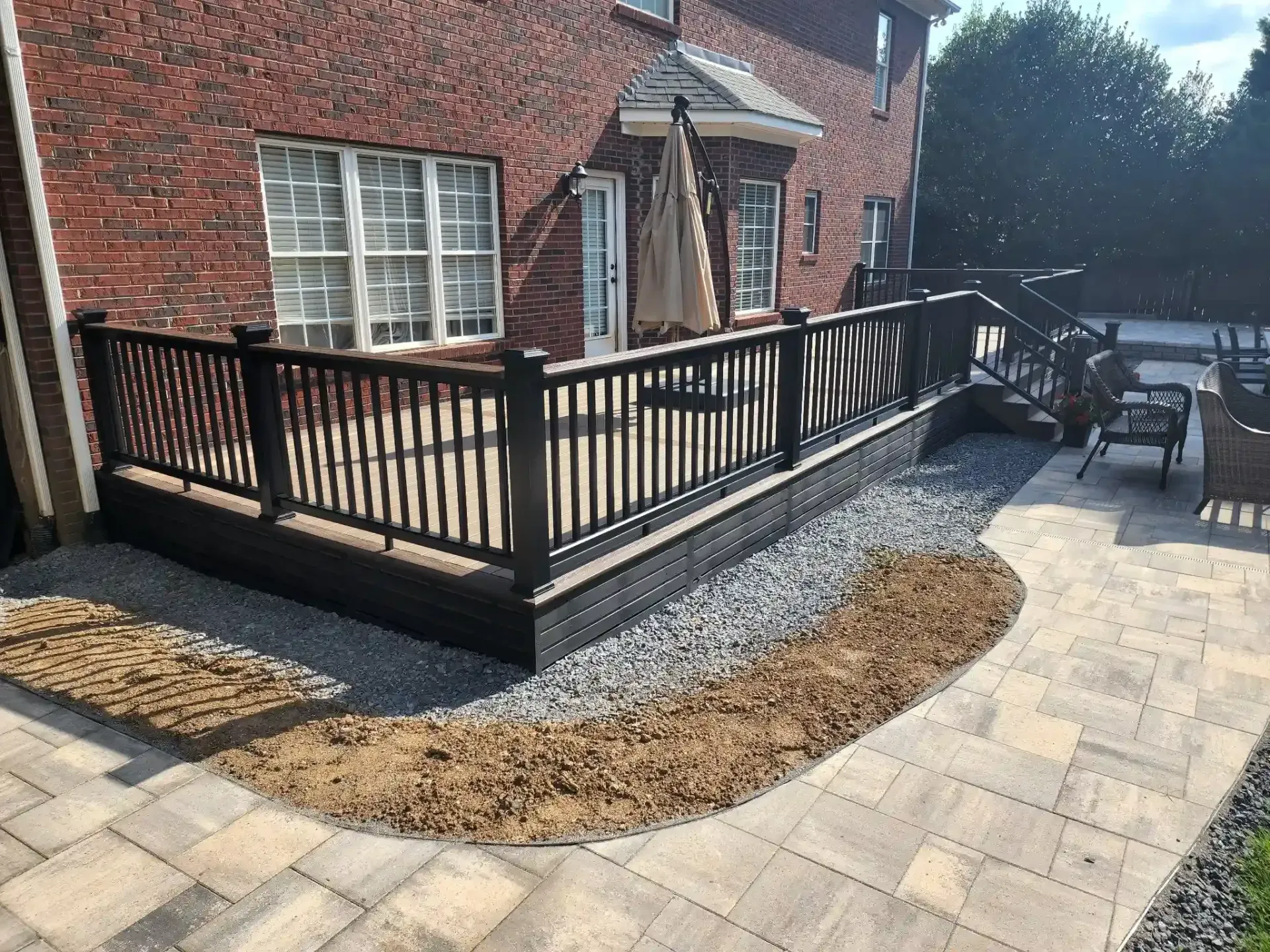 Dark brown deck and railing with gray gravel and mulch border, next to a brick building and paved patio.
