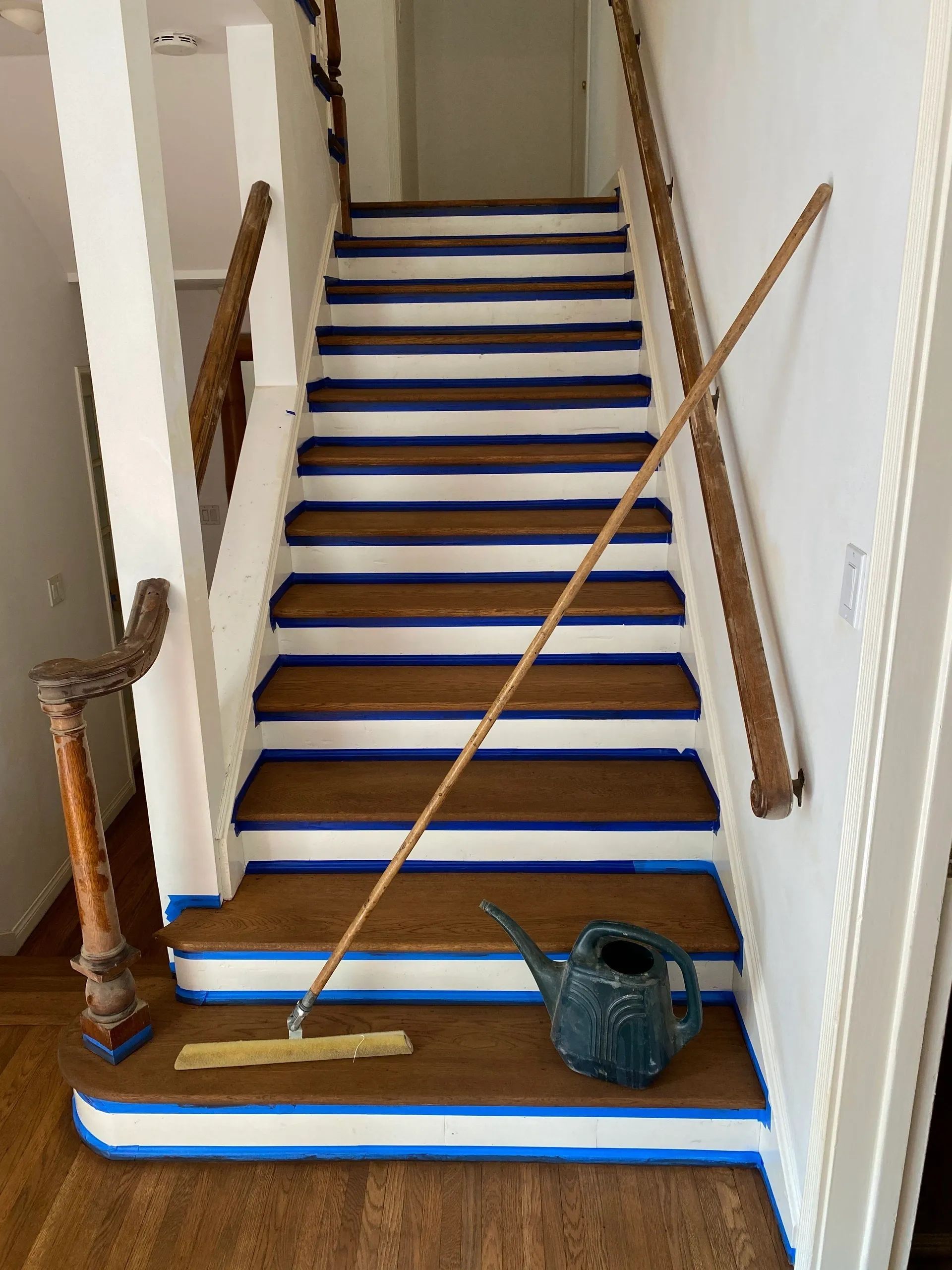 A mop is sitting on the bottom of a staircase next to a watering can