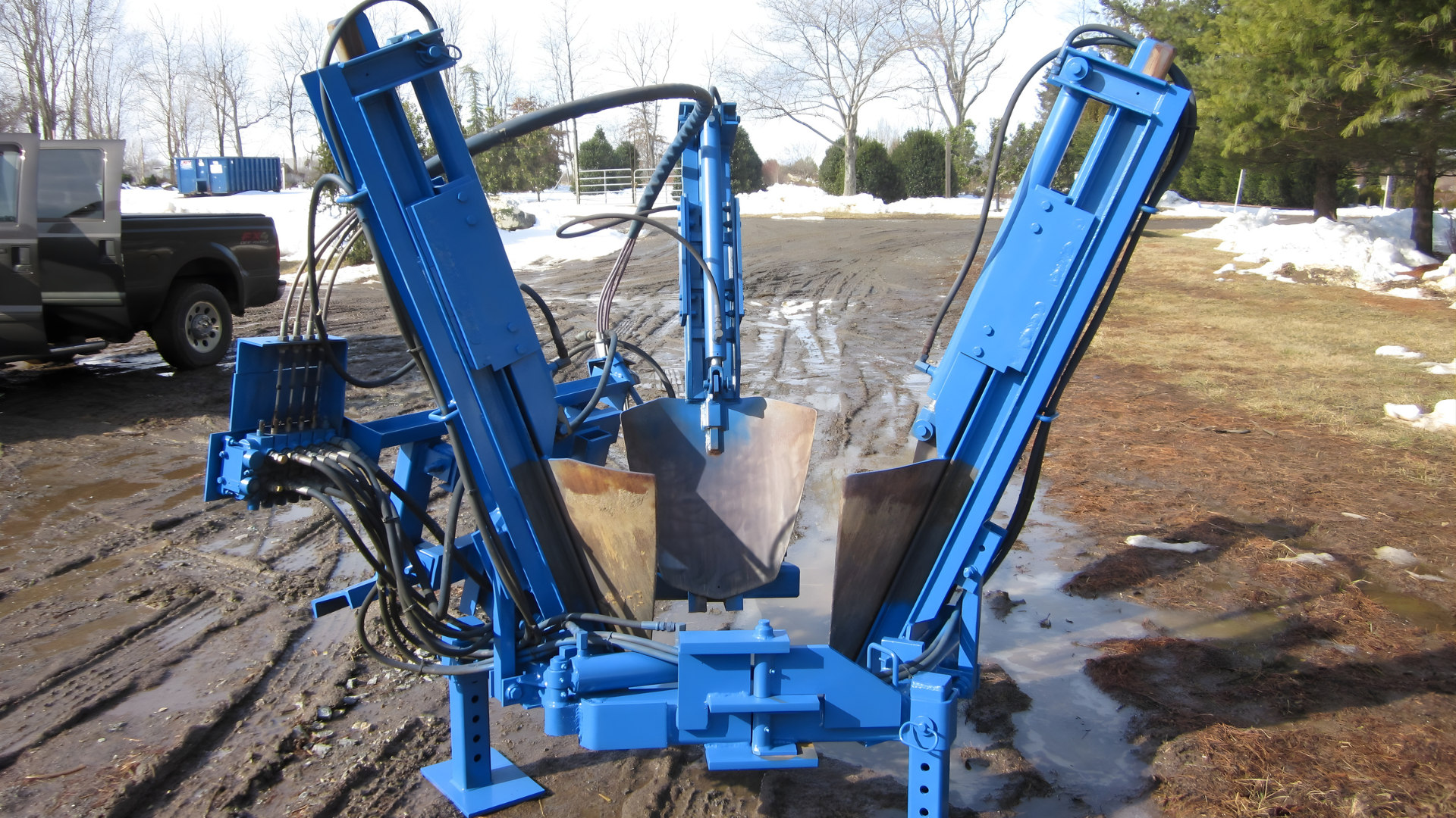 A bright blue hydraulic tree spade attachment stands on muddy, snowy ground next to a truck.