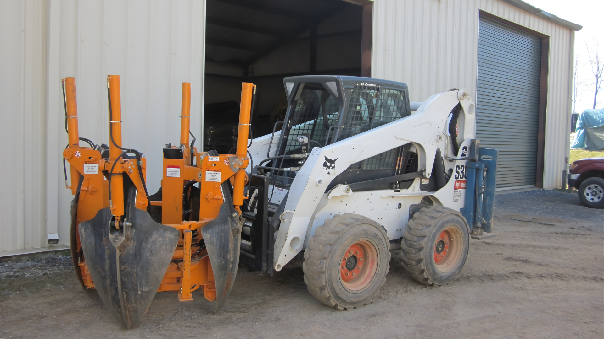 A white Bobcat skid-steer loader equipped with an orange tree spade attachment parked outside a metal building.