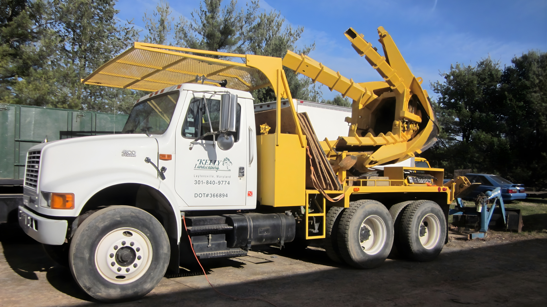 A white commercial truck with a yellow boom and mechanical arm assembly, parked outdoors on a dirt lot.