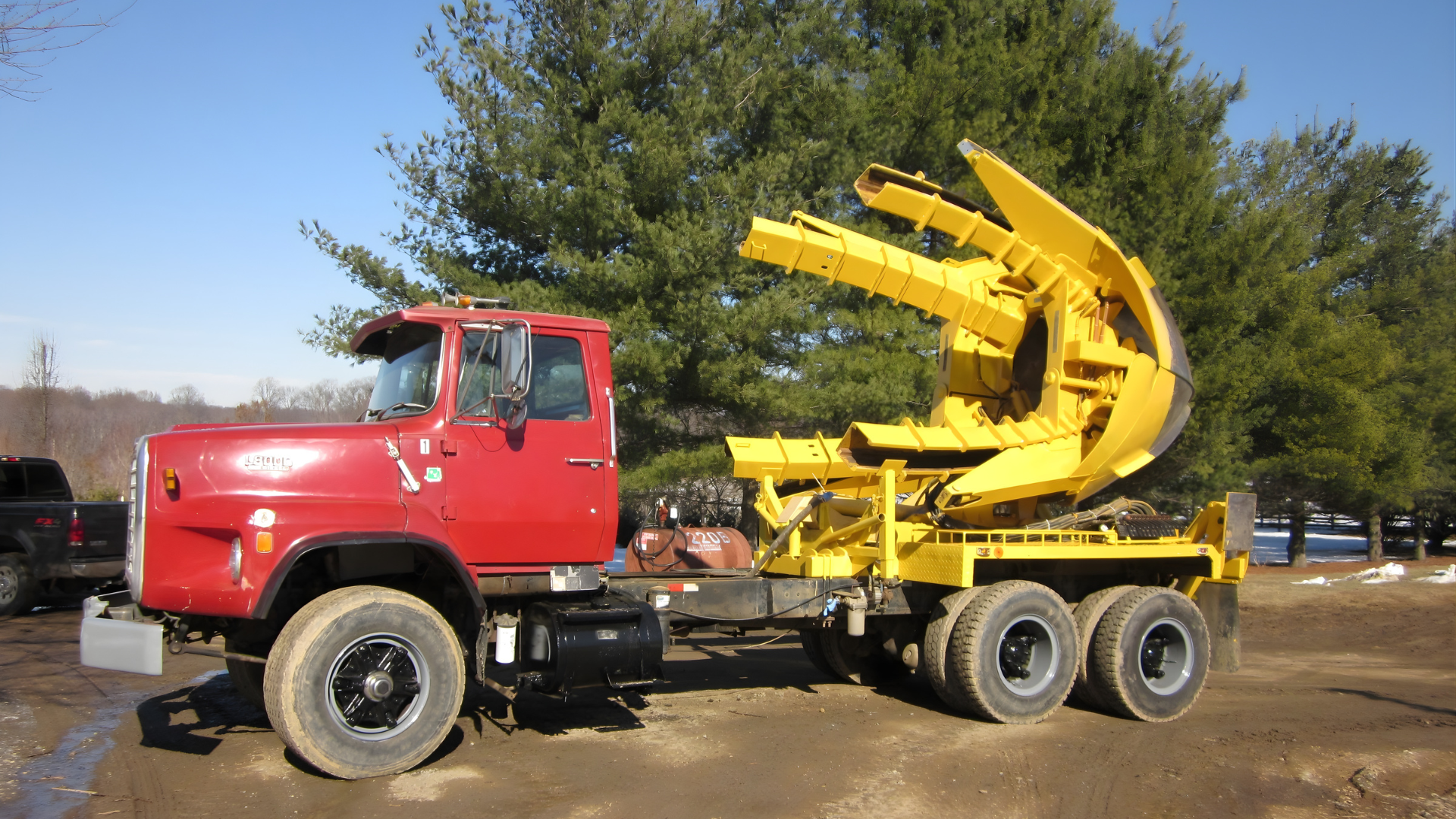 A red semi-truck with a specialized yellow tree-transplanting spade attachment on its bed parked outdoors.