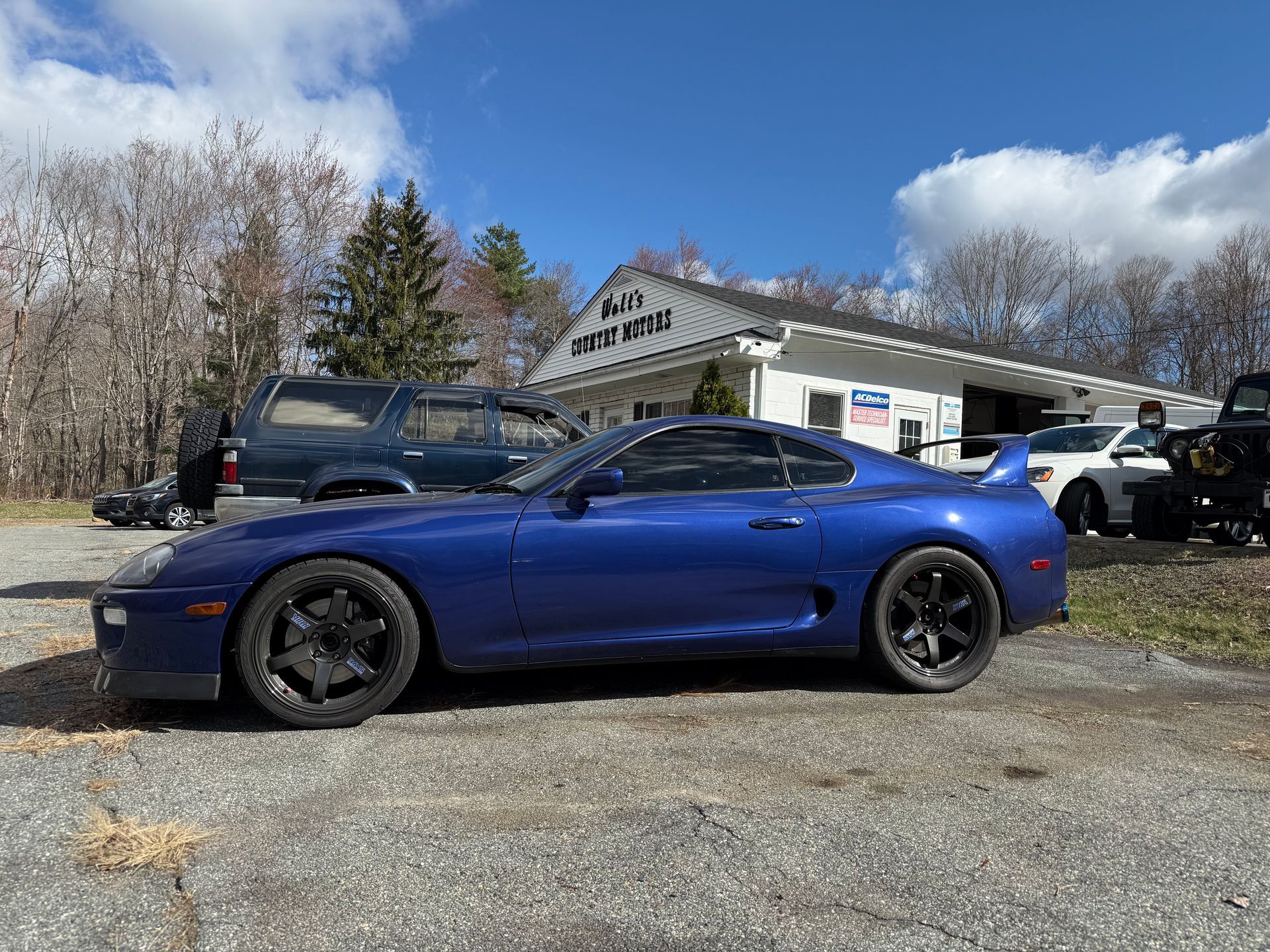 Blue sports car parked outside a small building on a gravel lot under a cloudy sky.