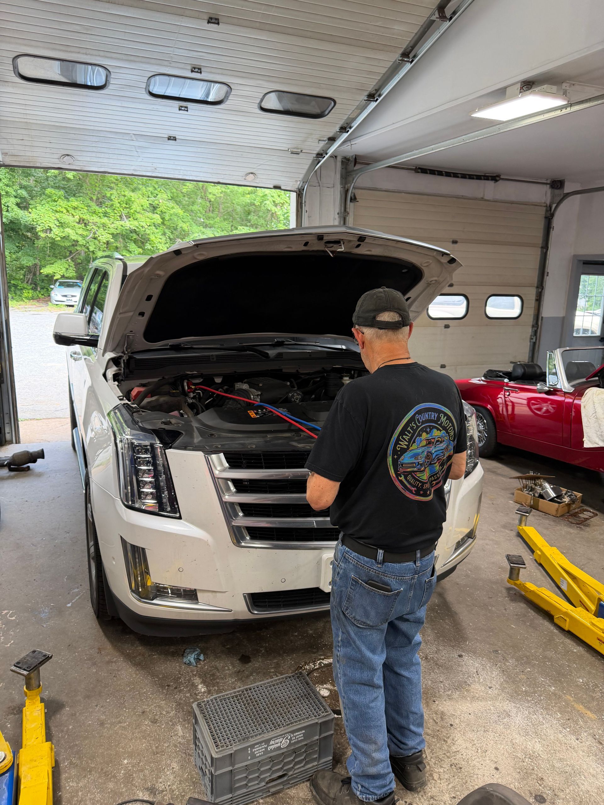 Mechanic inspecting a white SUV with open hood in a garage bay, tools and red car nearby