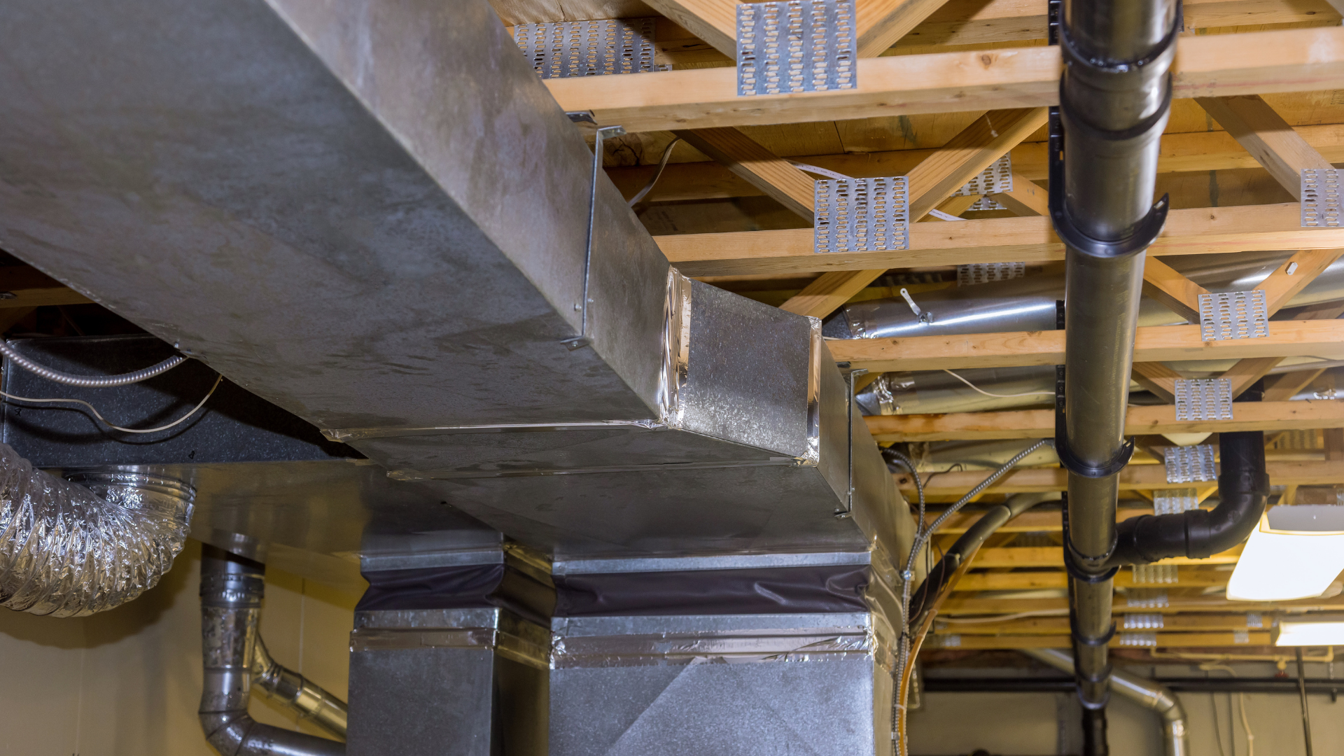 HVAC ductwork and plumbing pipes installed in a basement ceiling with wooden beams.