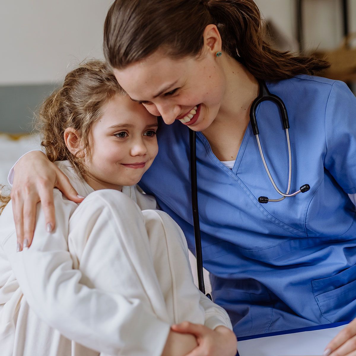 Smiling nurse in blue scrubs embracing a child in a white robe in a warm clinic setting