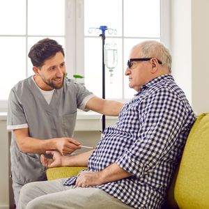 Caregiver in gray scrubs gently comforts a seated man in a plaid shirt beside an IV stand Caregiver in gray scrubs gently comforts a seated man in a plaid shirt beside an IV stand
