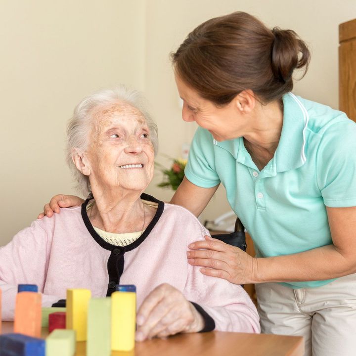 Caregiver in teal shirt gently comforts an elderly woman at a table with colorful blocks in a home setting Caregiver in teal shirt gently comforts an elderly woman at a table with colorful blocks in a home setting