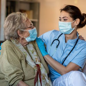 Nurse in blue scrubs and mask comforting a masked elderly patient indoors Nurse in blue scrubs and mask comforting a masked elderly patient indoors