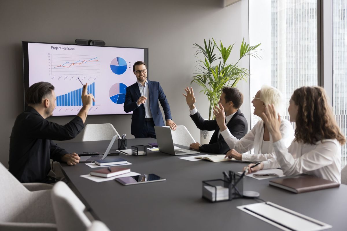 Business meeting: man presenting, colleagues raising hands, graphs on screen. Modern office.