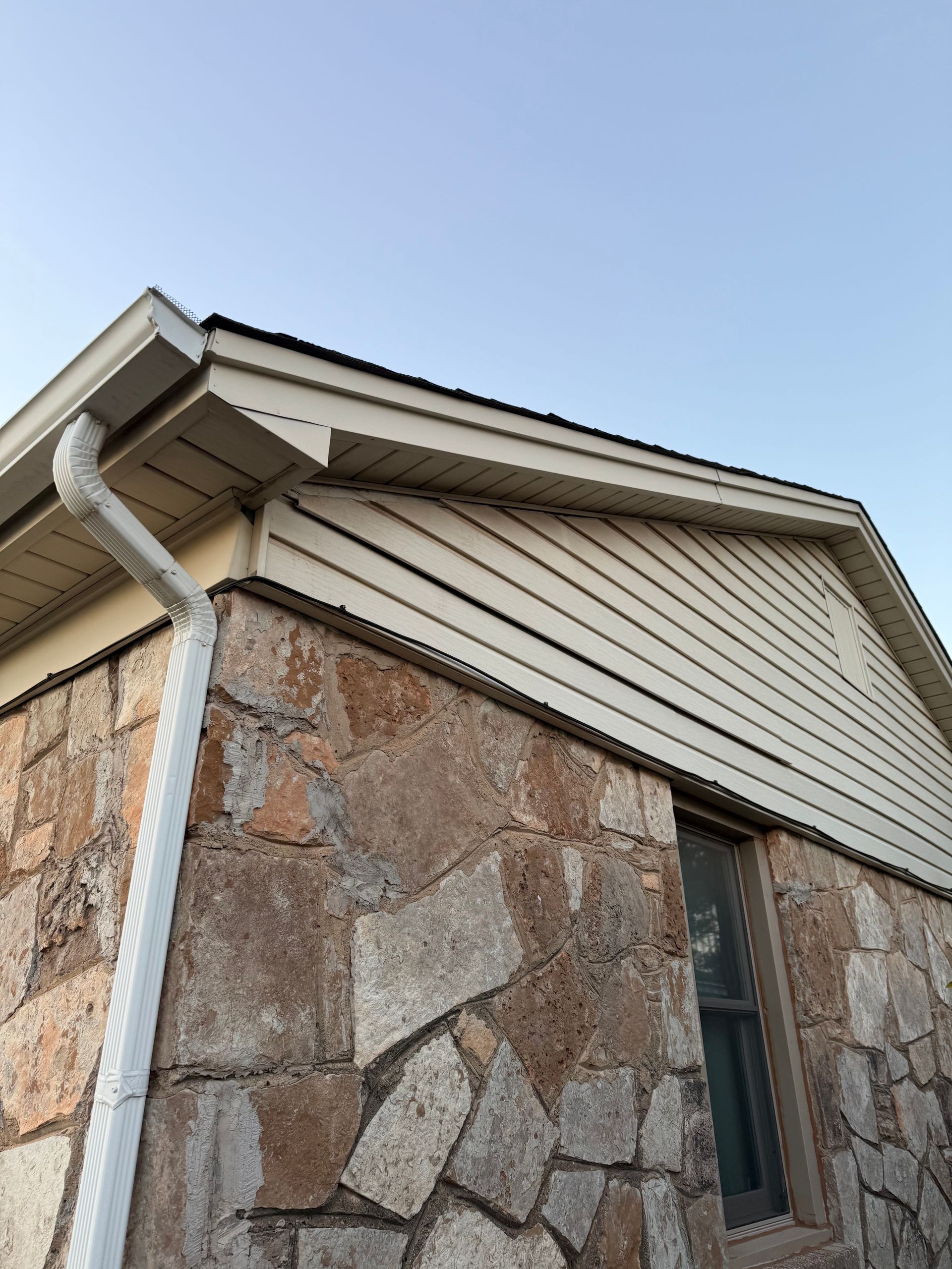 Stone-walled building with light siding and white gutter under a clear blue sky.