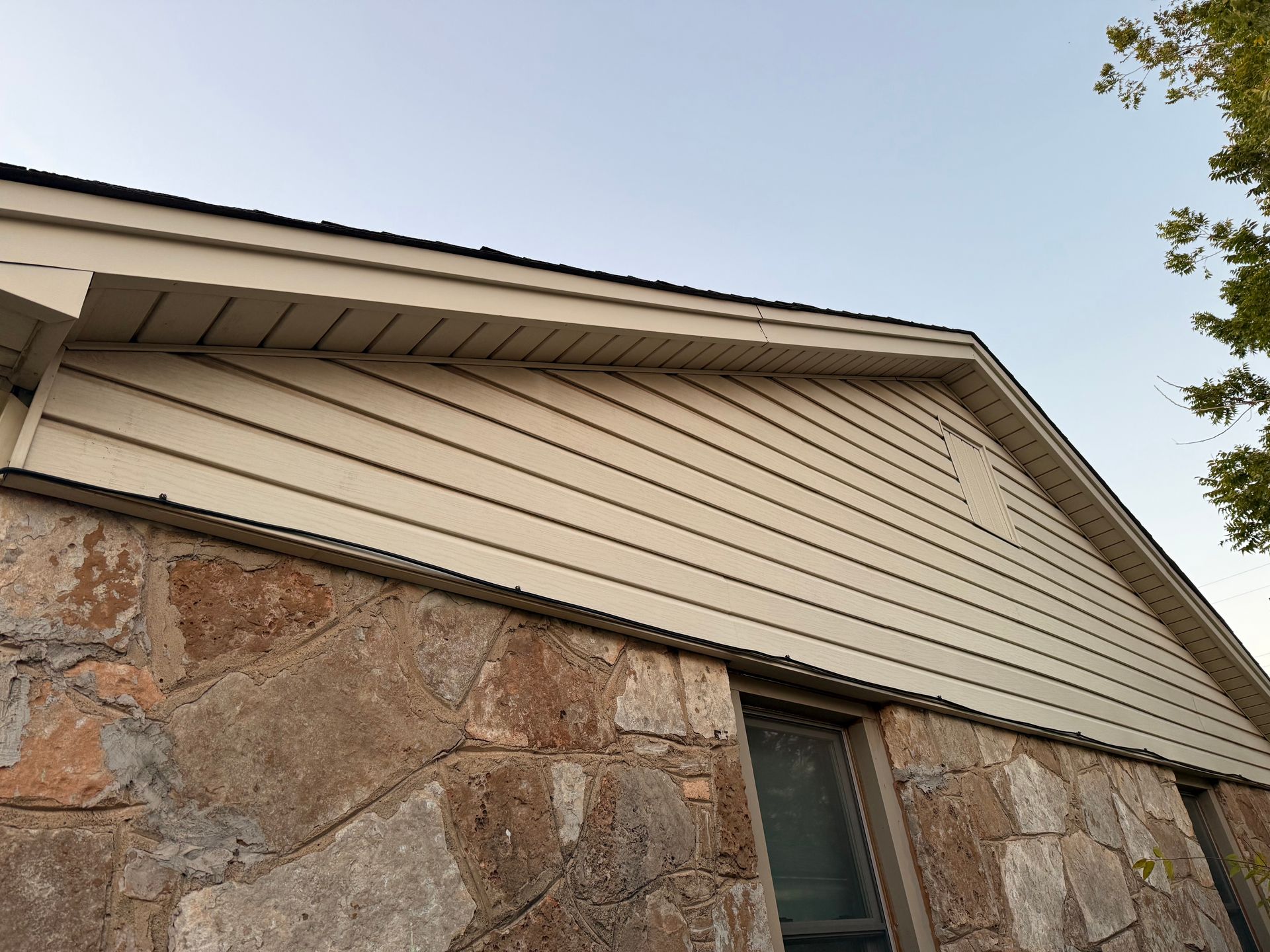 Stone exterior of a building with light beige siding beneath the roofline; a window is visible.