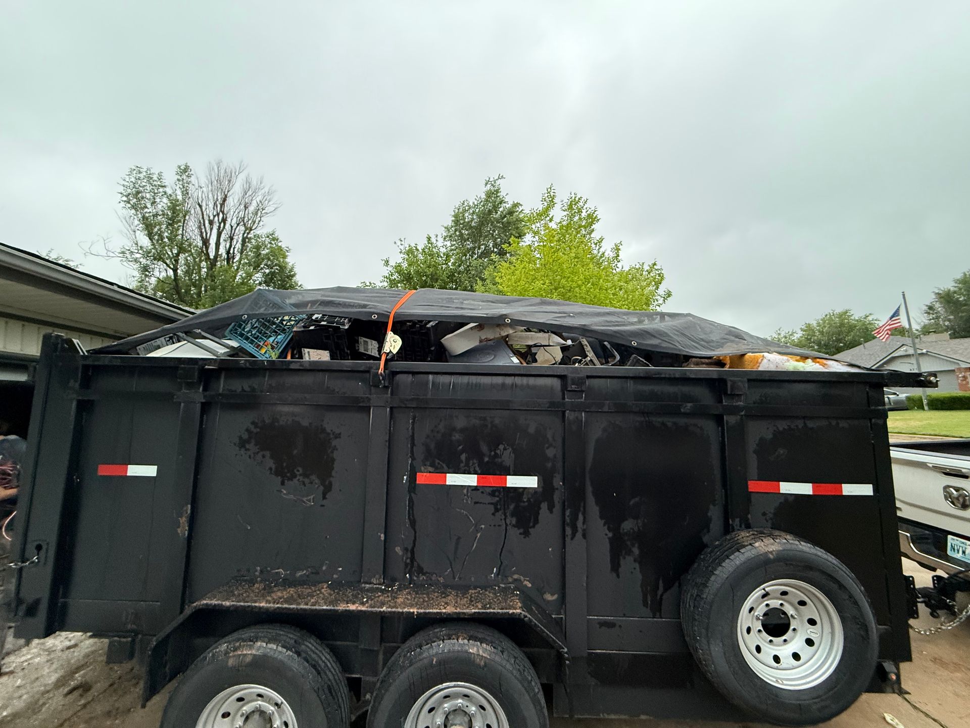 Black trailer filled with debris, covered with a tarp secured by an orange strap, under an overcast sky.
